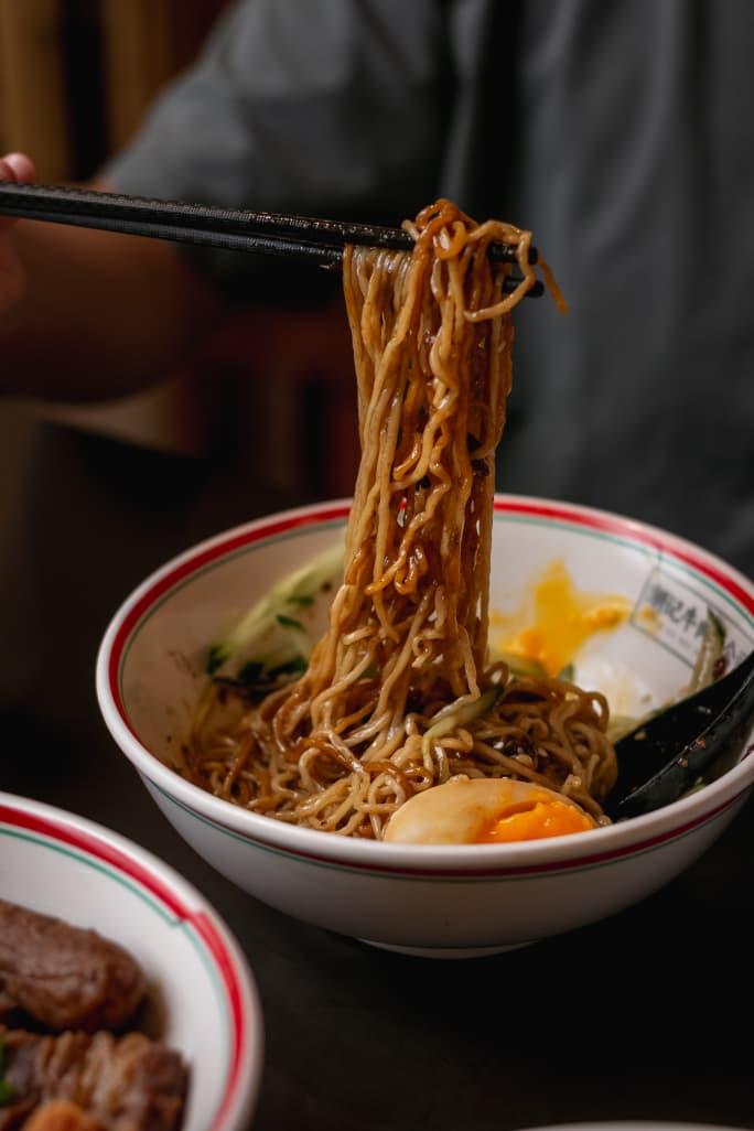 Chopsticks lifting dark-sauce dry tossed noodles from a Teowkee bowl