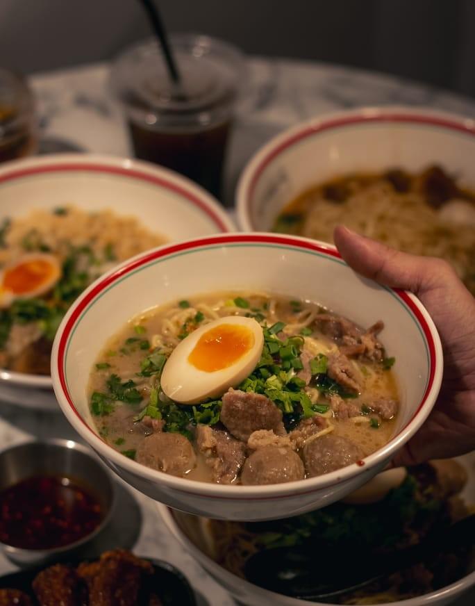 Hand holding a Teowkee beef noodle soup bowl with soft-boiled egg and meatballs
