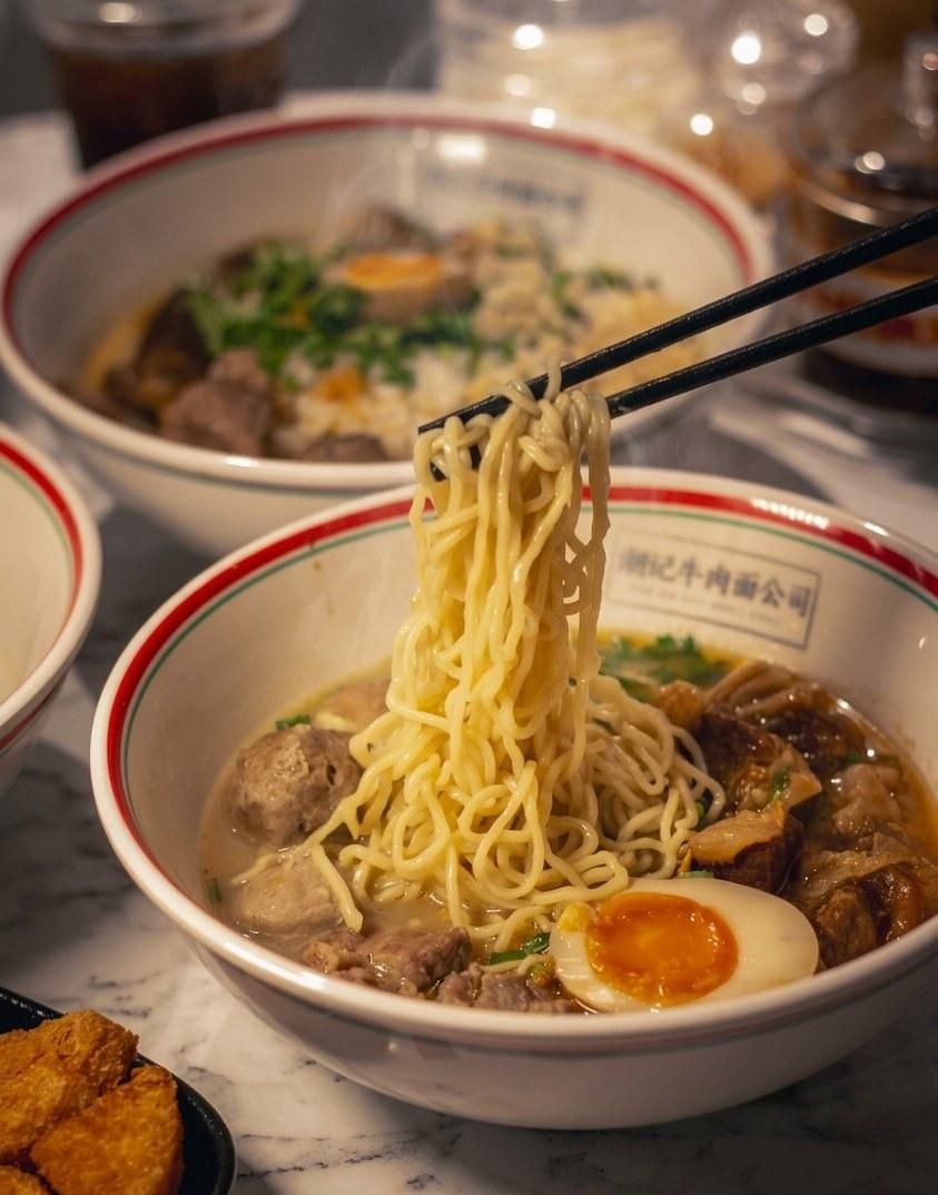 Chopsticks lifting noodles from a steaming Teowkee beef noodle bowl with soft-boiled egg